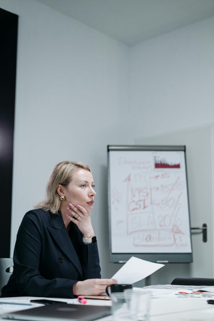 A businesswoman in a black suit analyzing a document in an office setting with a presentation board in the background.